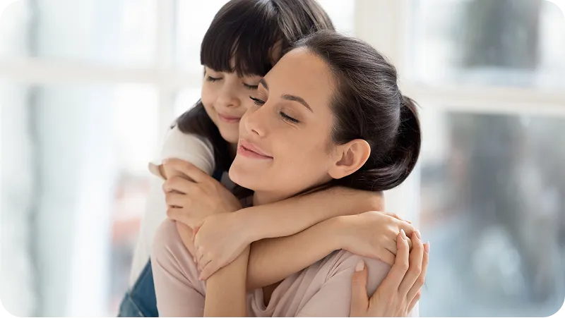 A young girl with dark hair hugging her mother, both smiling with eyes closed.
