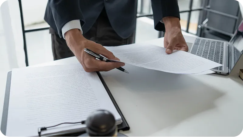A person in a suit stands over a desk, holding a pen and reviewing or organizing several legal documents.