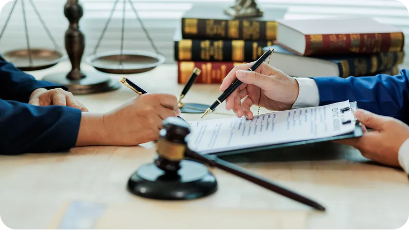 A legal desk where two people are signing a contract document, with a gavel and scales of justice.