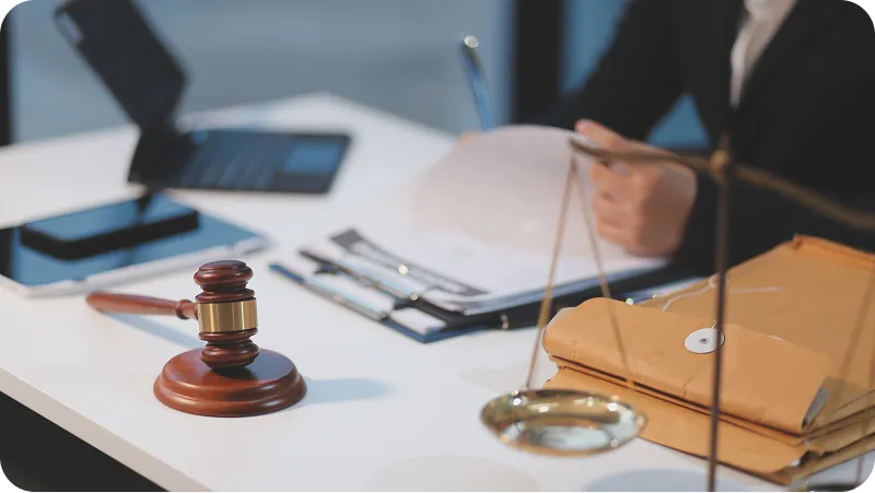A legal professional is seen signing papers on a white desk, with a gavel and scales of justice on it.