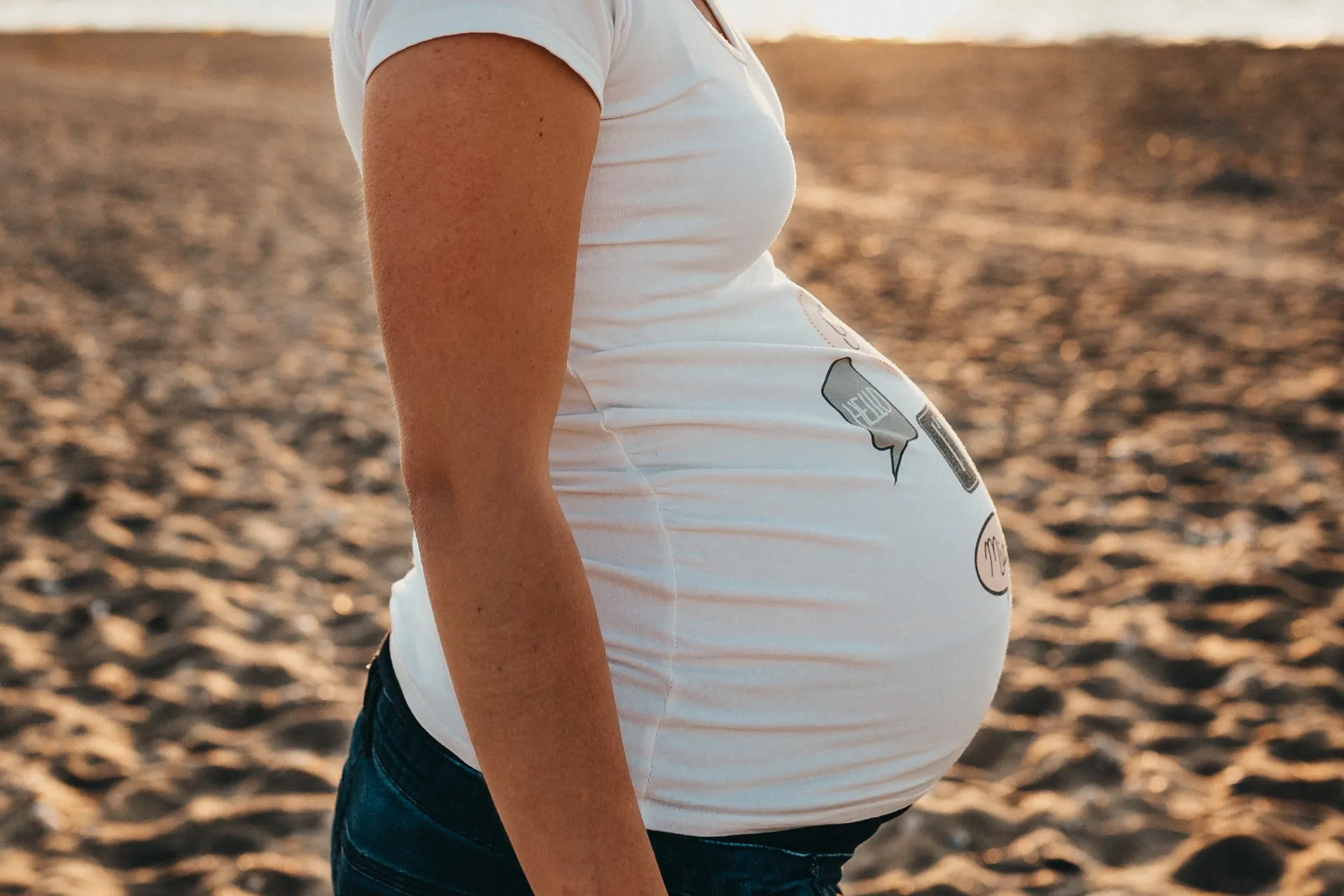 A pregnant woman in a side view, facing to her right, standing on the sand near the sea.