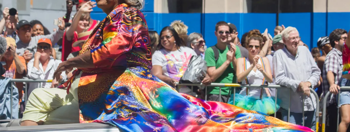 A man wearing a colorful long dress, with many people in the background watching him.