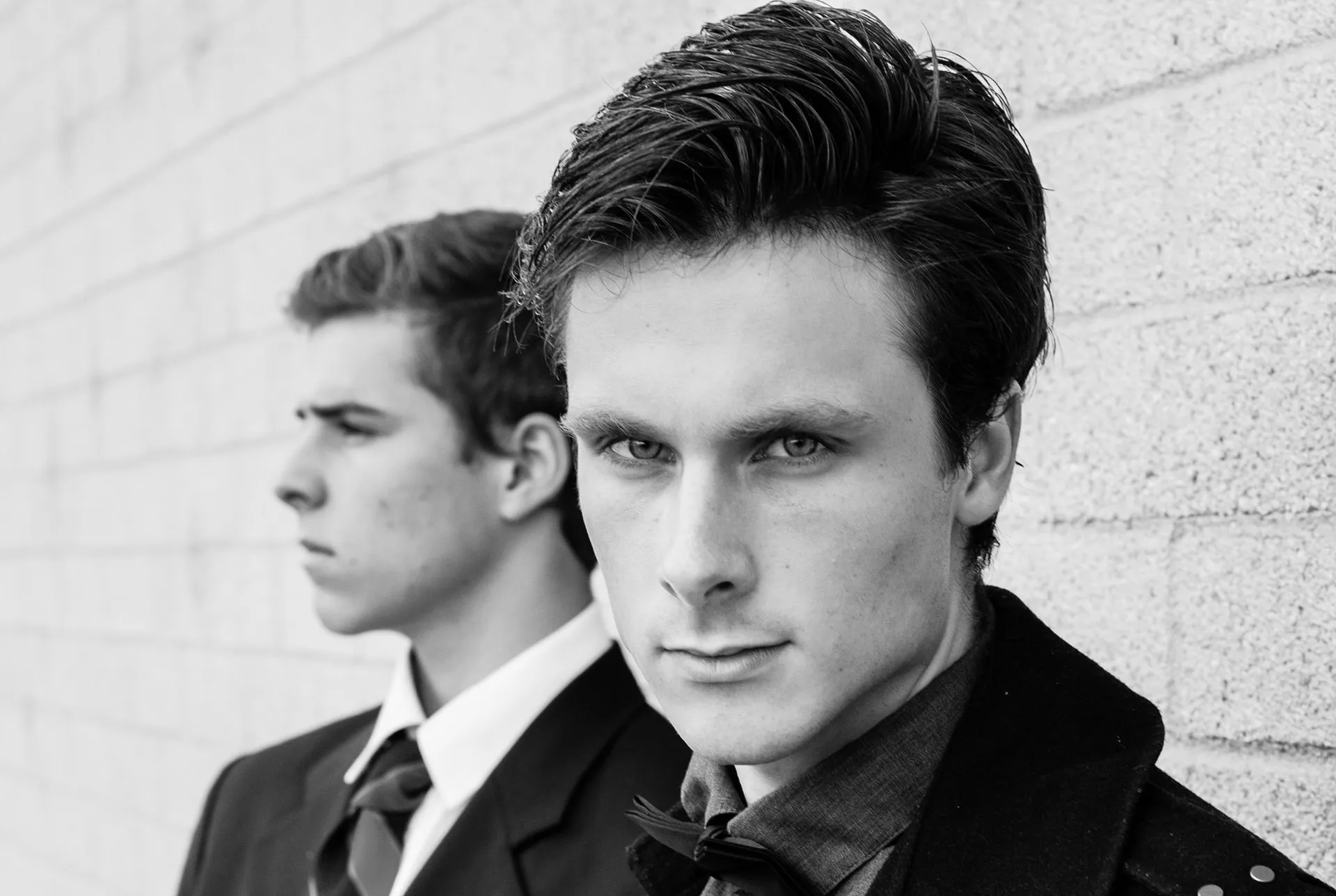 Two young men in formal attire stand against a textured wall in a black and white photograph.