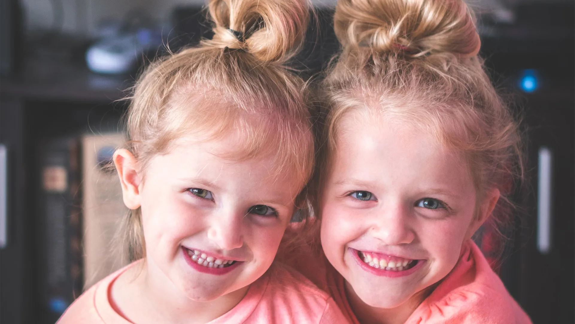 An image of two smiling, beautiful baby girls wearing the same color tops and having the same hairstyle.