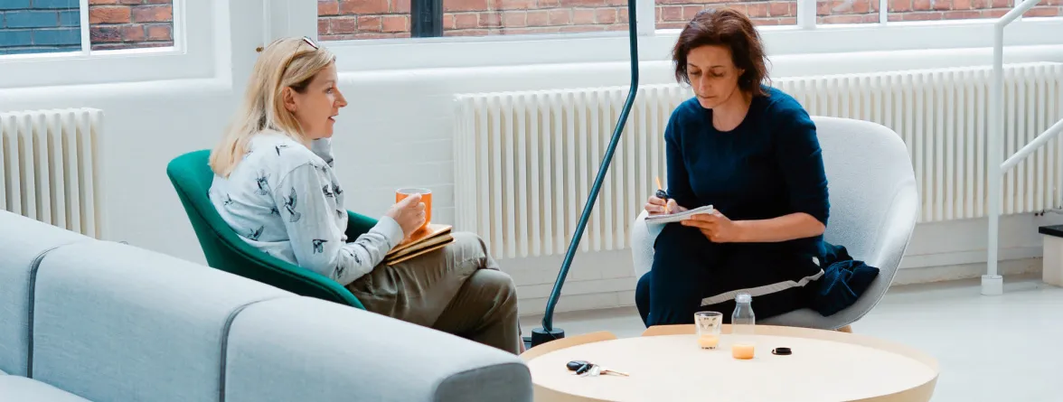 Two women sitting facing each other, with one woman taking information and writing it down in her notes while the other woman holds a cup of coffee.