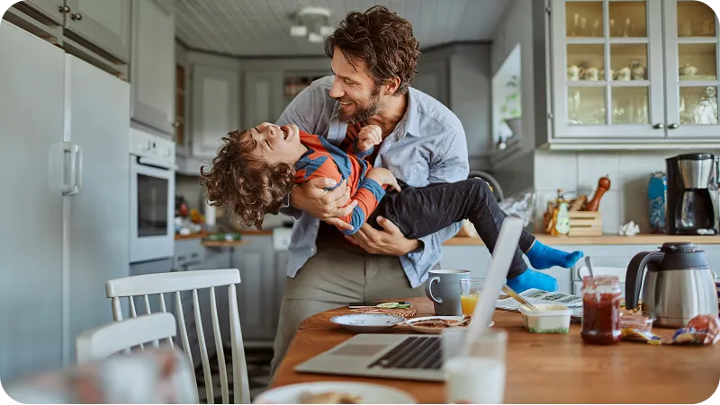 A father in a kitchen is happily lifting and playing with his laughing young son.