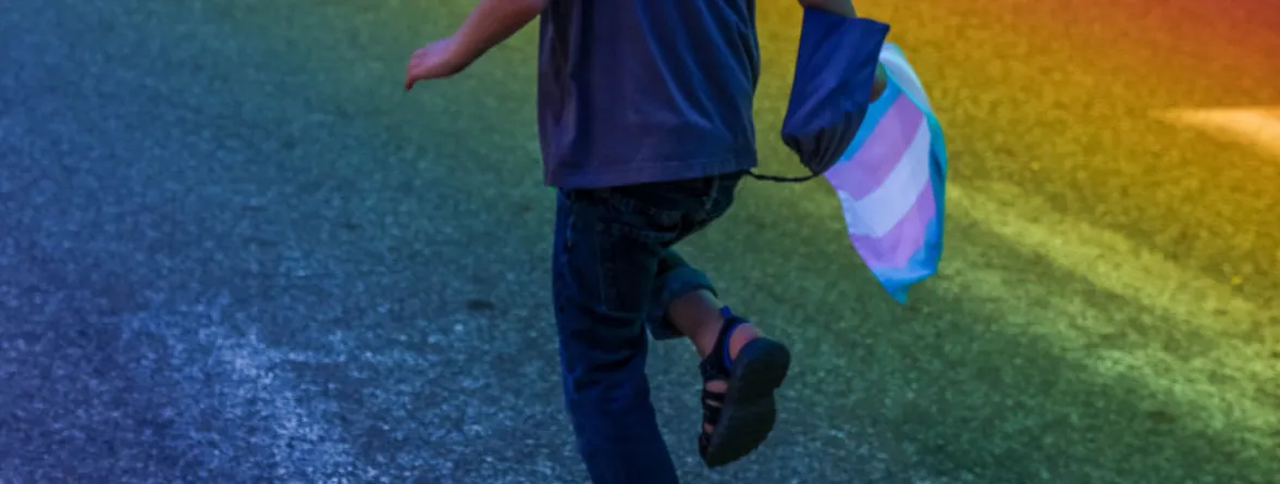 "A man walks against a rainbow-colored background, holding a flag in his right arm.